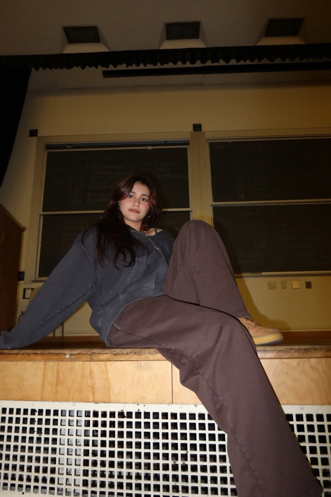 Student in a blank grey zip hoodie and brown sweatpants seated on a lecture hall ledge