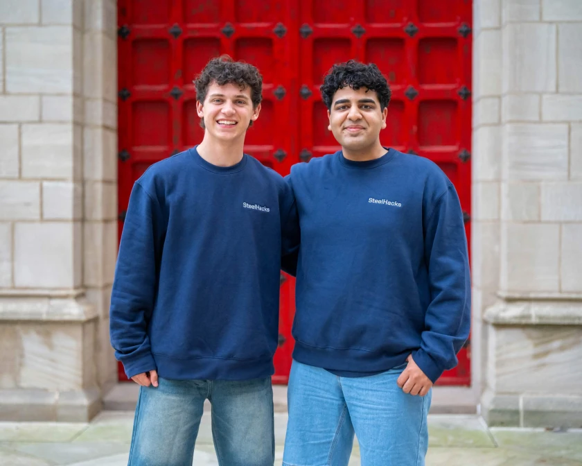 Two SteelHacks XIII participants in navy crewnecks and jeans in front of red doors