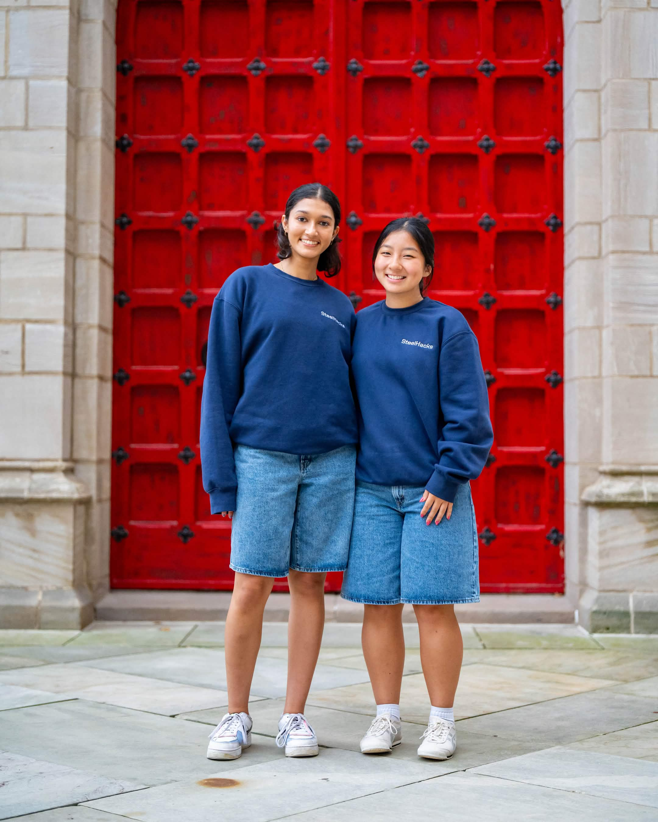 Two SteelHacks XIII participants in navy crewnecks and denim shorts in front of red doors