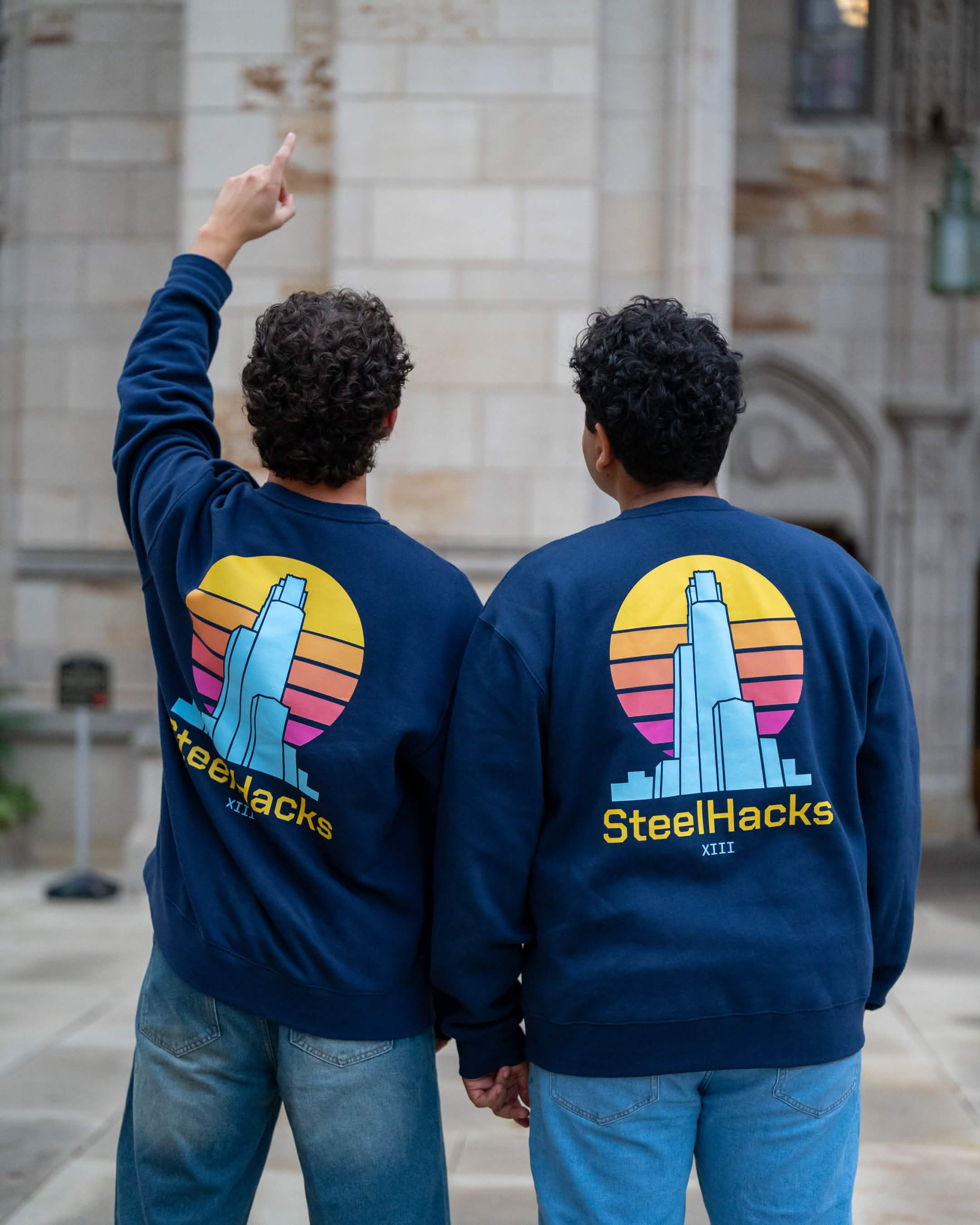 Two participants wearing navy SteelHacks XIII crewnecks from the rear, showing the sunset Pittsburgh skyline back graphic