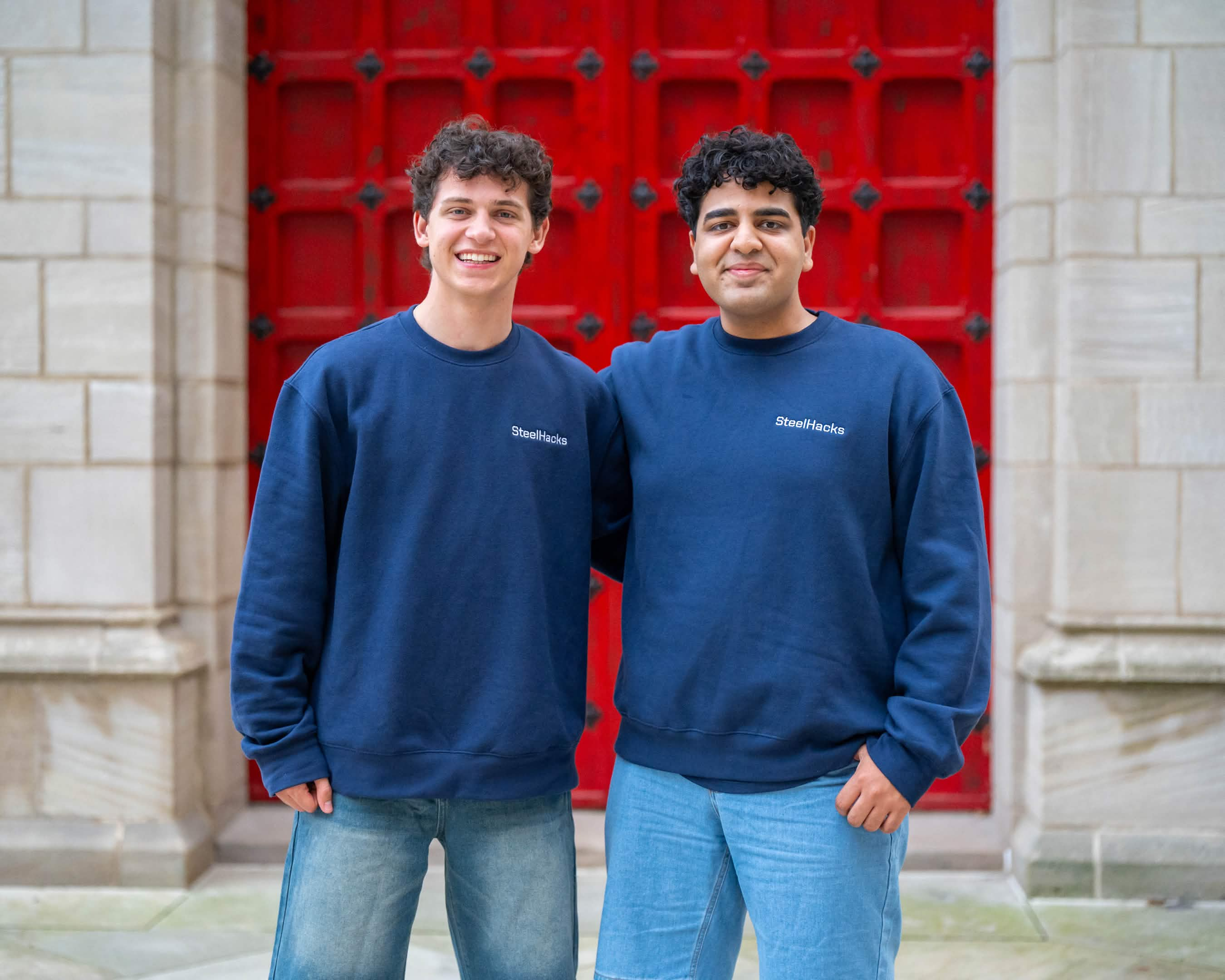 Two SteelHacks XIII participants in navy crewnecks and jeans in front of red doors